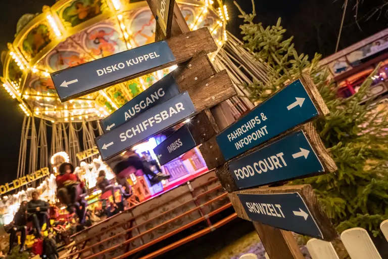 Hölzerne Wegweiser auf dem Gelände zeigen zu Food Court, Showzelt, Schiffsbar und Disko Kuppel, dahinter dreht sich ein beleuchtetes Karussell