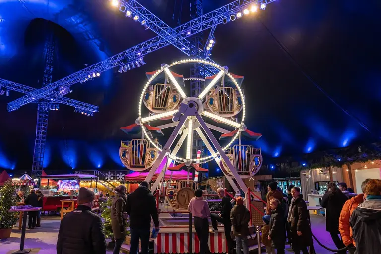 Beleuchtetes Riesenrad unter dem Lichterdach des Kuppelzelts, davor bummeln Besucher zwischen Karussell und Buden