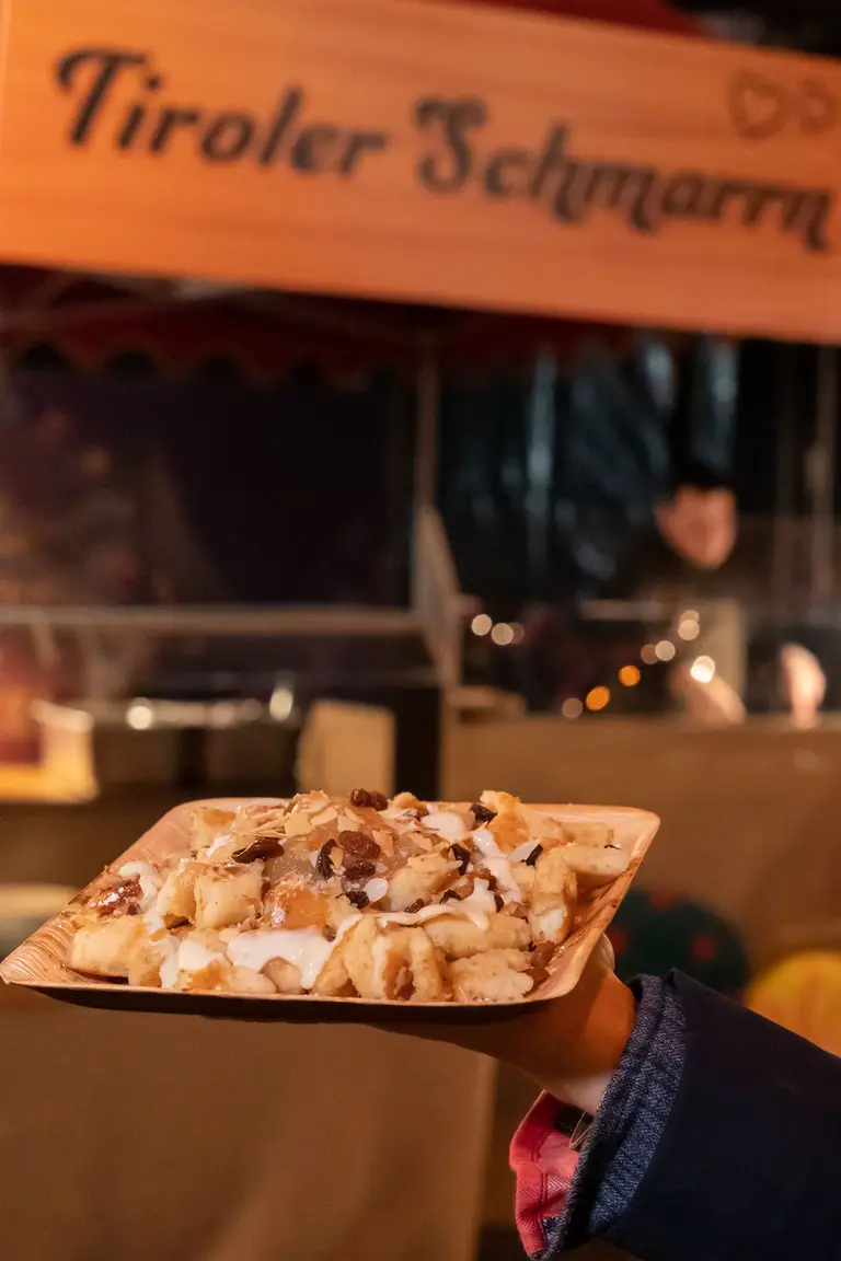 Hand hält eine Schale frischen Kaiserschmarrn mit Mandeln vor dem warm beleuchteten Stand Tiroler Schmarrn