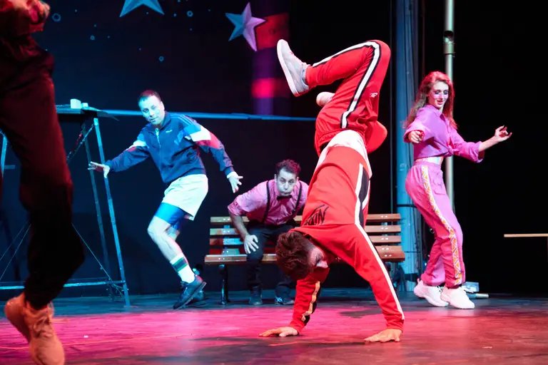 Breakdancer im roten Trainingsanzug steht im Handstand auf der Bühne, umringt vom Ensemble