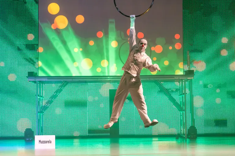 Clown in weißem Kostüm mit roter Nase springt über die Bühne und greift nach einem Ring